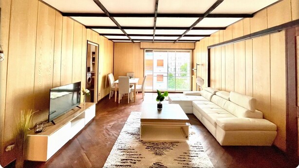 A modern living room with a sofa, a coffee table, and a TV. There is a dining area with chairs and a table. The room has wooden walls and a ceiling with a grid pattern. - Family Friendly 2BR Apartment • Terrace - Parking - Neuilly-sur-Seine/La Defense (Neuilly-sur-Seine)