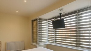 A bedroom with a large bed, a bedside table, a radiator, and a window with blinds. - The Miles Arms Hotel (Bristol)