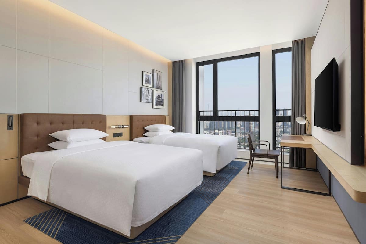 Habitación de hotel con dos camas, un escritorio, una silla, un televisor y ventanas amplias con vistas a la ciudad.