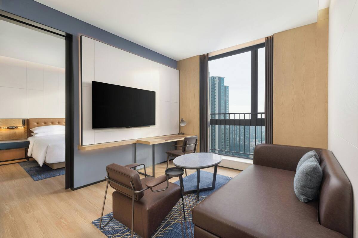 Una habitación de hotel moderna con una cama, un televisor de pantalla plana en la pared, un rincón de comedor con mesa y sillas, un sofá y un amplio ventanal con vistas a la ciudad.