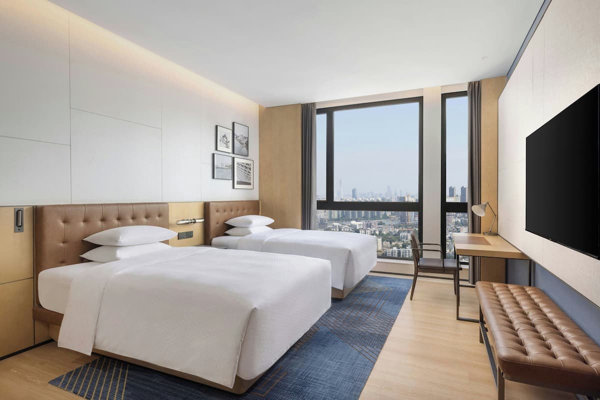 Habitación de hotel con dos camas, un ventanal grande con vista a la ciudad, un escritorio con una silla y un televisor de pantalla plana.