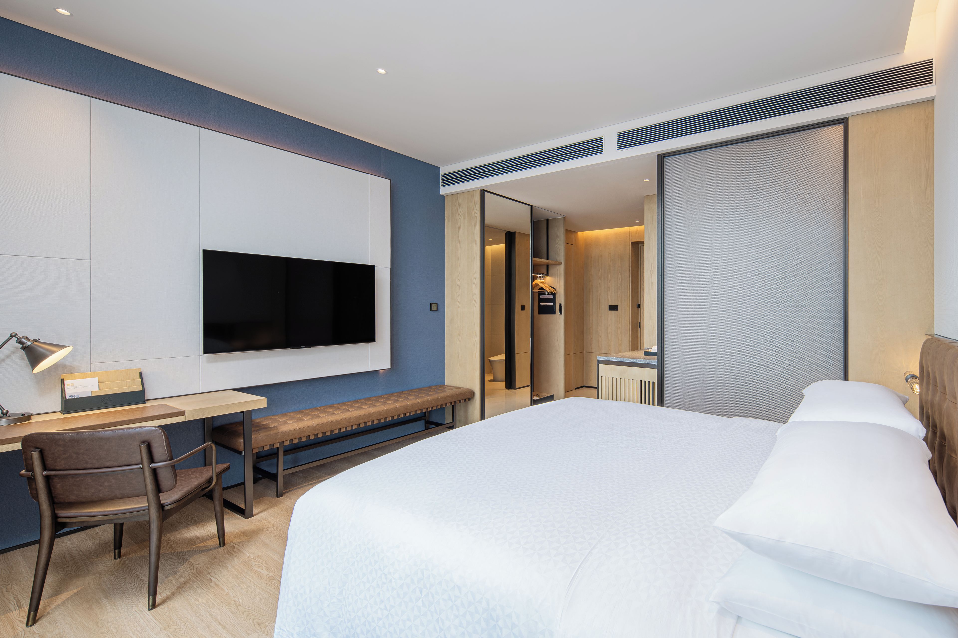 Una habitación de hotel moderna con una cama grande, un escritorio con silla, televisión y baño visible a través de una puerta corredera.