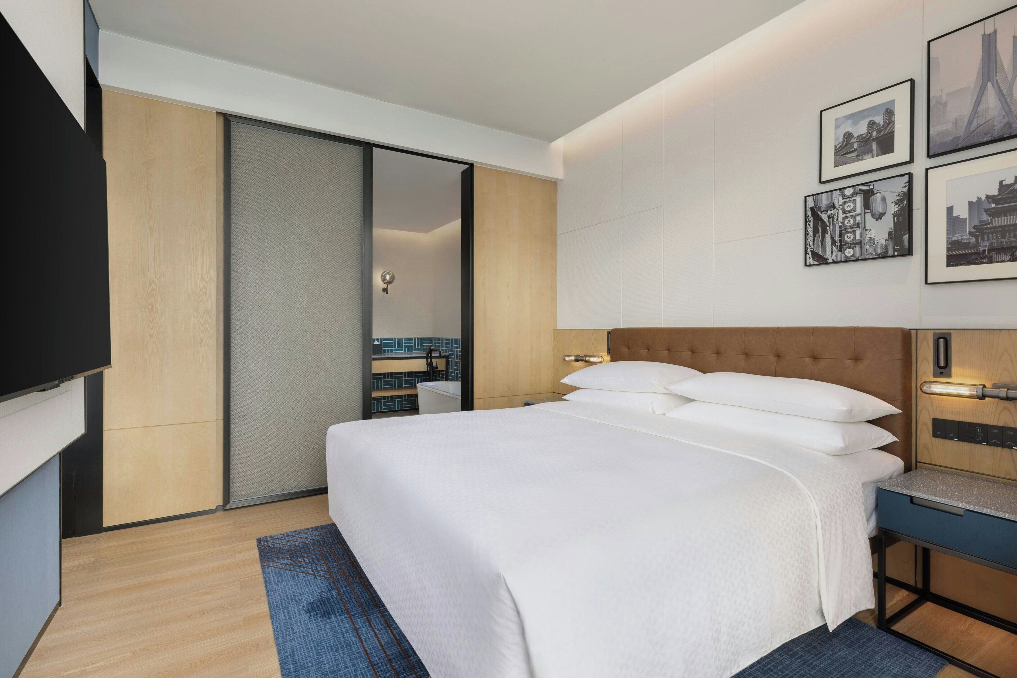 Ein modernes Hotelzimmer mit einem großen Bett, einem Flachbildfernseher, einem Holz-Kopfteil und einem an der Wand befestigten Kunstwerk.