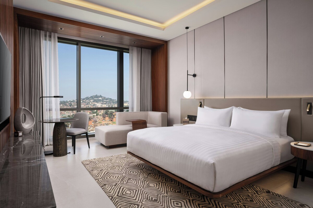Een moderne hotelkamer met een groot bed, een zithoek met een bank en fauteuil, een bureau met stoel, en uitzicht op de stad door grote ramen.