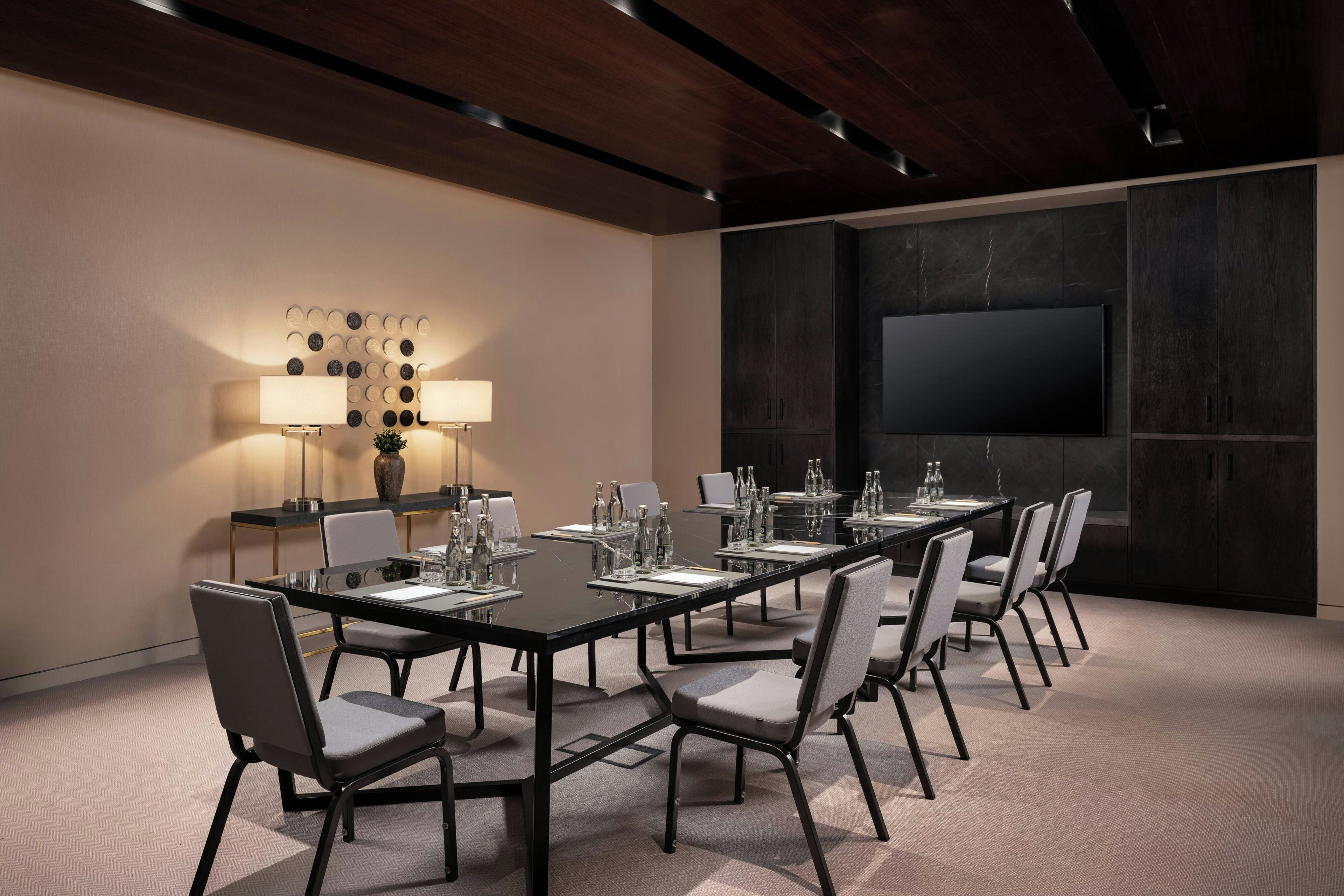 A conference room with a long table, chairs, a TV screen, and wall-mounted lamps.
