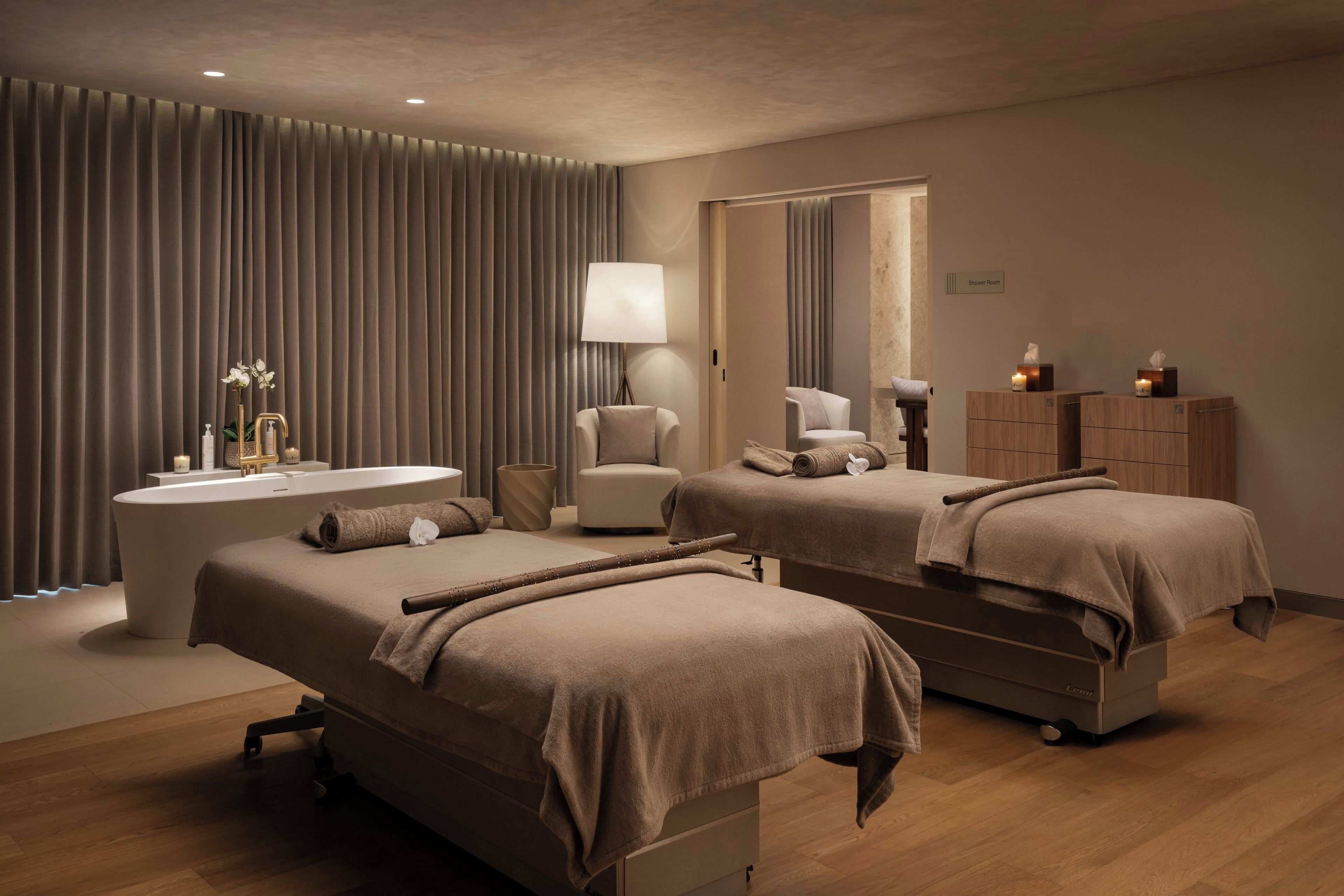 A hotel room with two massage beds, a bathtub, a vanity with a mirror, and a shower area.