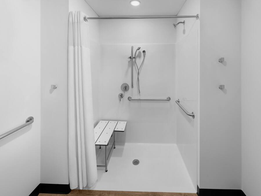 A modern shower room with a white tiled wall, a showerhead, a handheld shower, a handheld showerhead, a grab bar, and a small table with a towel.