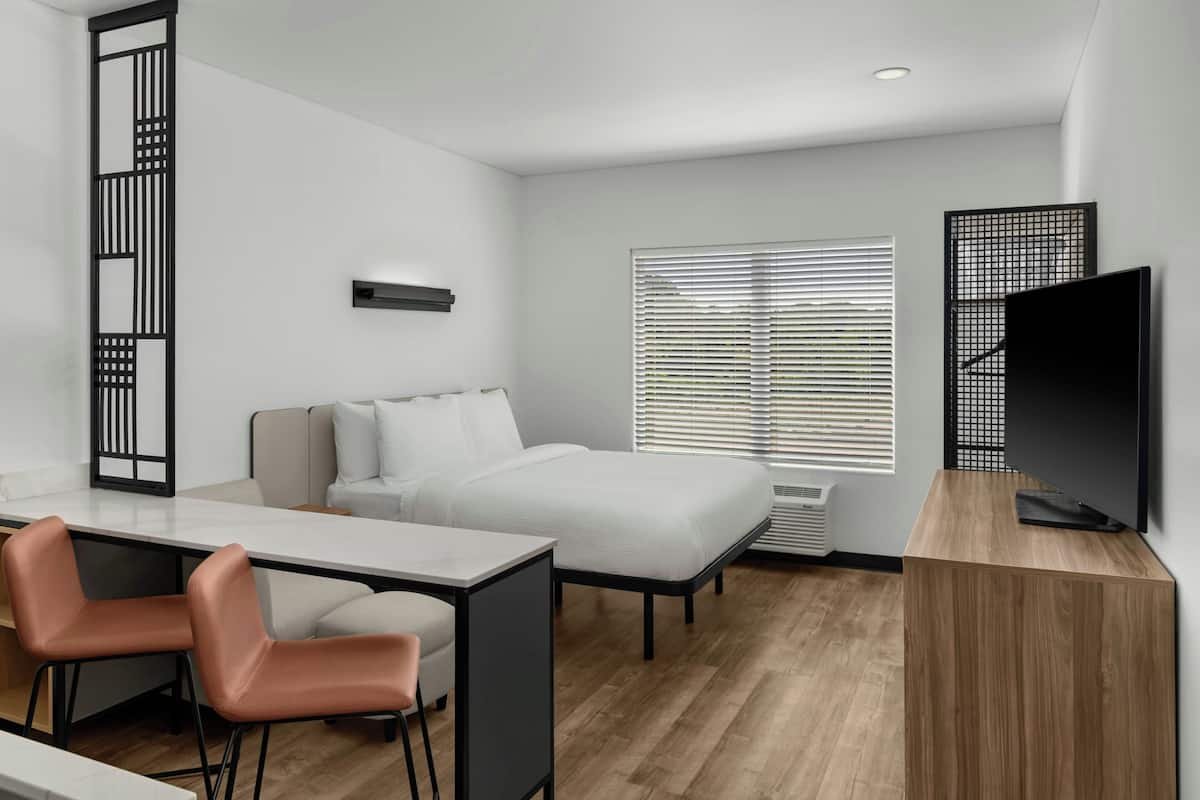A modern hotel room with a bed, a desk with chairs, a television, and a window with blinds.