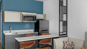 A compact kitchen with a microwave, refrigerator, and sink. There is a dining table with two chairs and a sofa with a patterned pillow. - StudioRes by Marriott Greensboro Airport (Greensboro)
