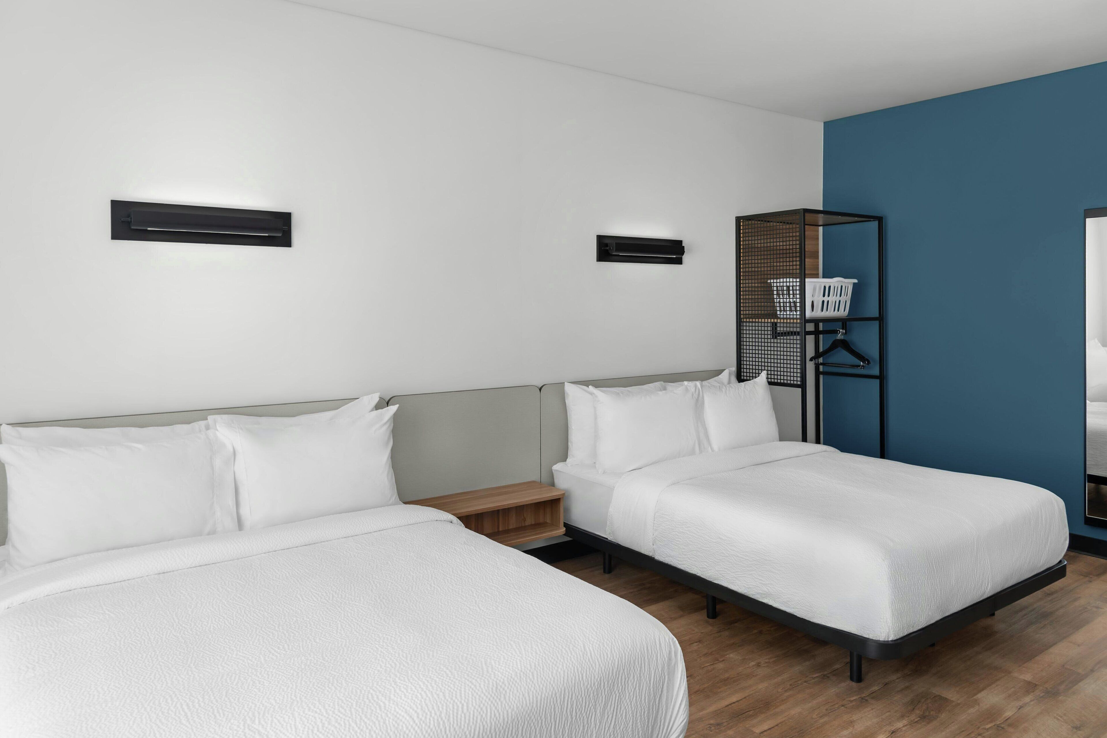 Une chambre d’hôtel avec deux lits, une table de chevet en bois, une étagère murale avec un panier, et un mur d’accent bleu.