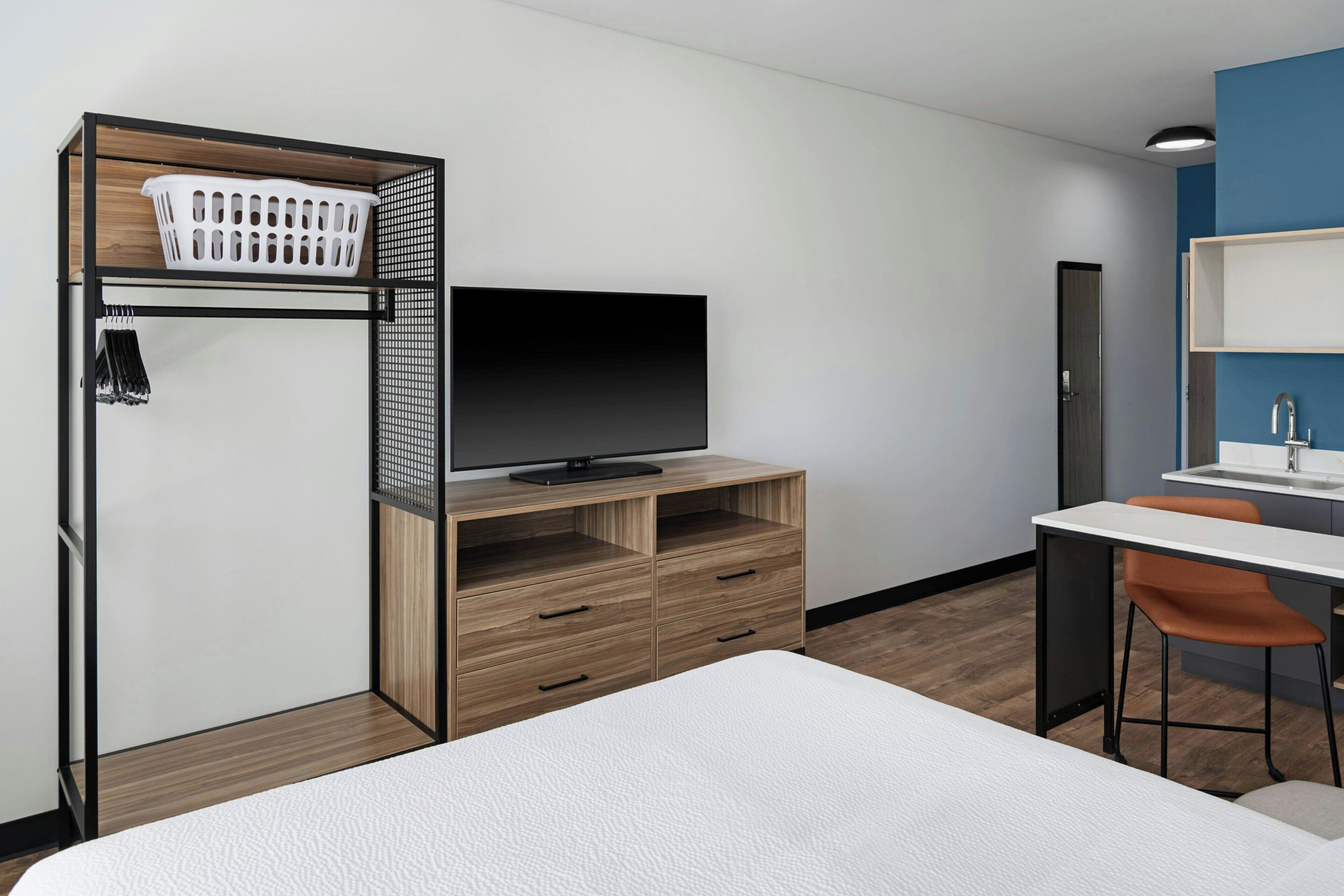 Una habitación de hotel moderna con cama, televisor sobre un soporte de madera, escritorio con silla y baño con lavabo y espejo.
