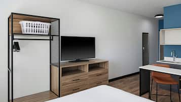 Una habitación de hotel moderna con cama, televisor sobre un soporte de madera, escritorio con silla y baño con lavabo y espejo.