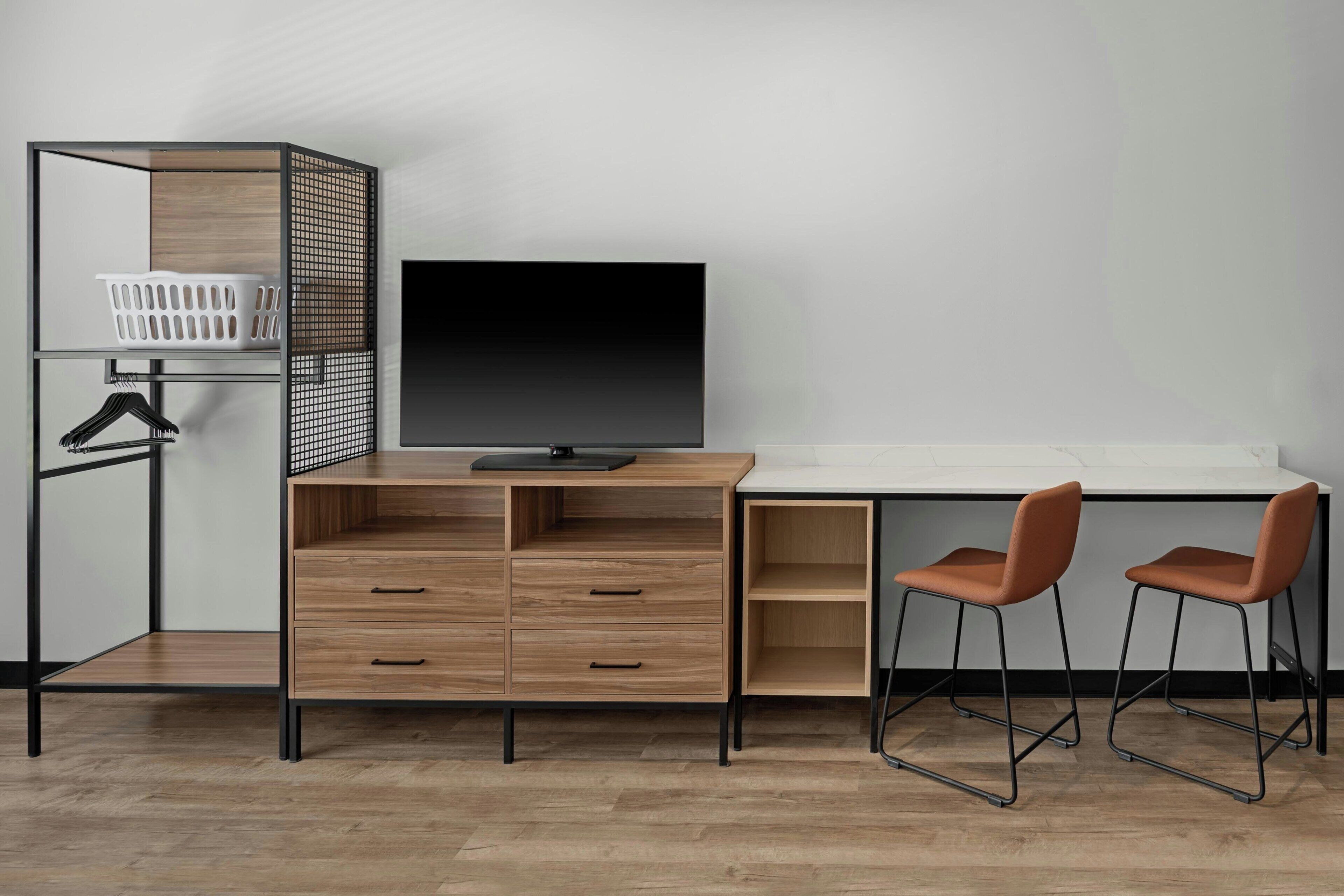 Habitació moderna amb escriptori, dues cadires, televisió, cistella de roba i una prestatgeria per a la roba.