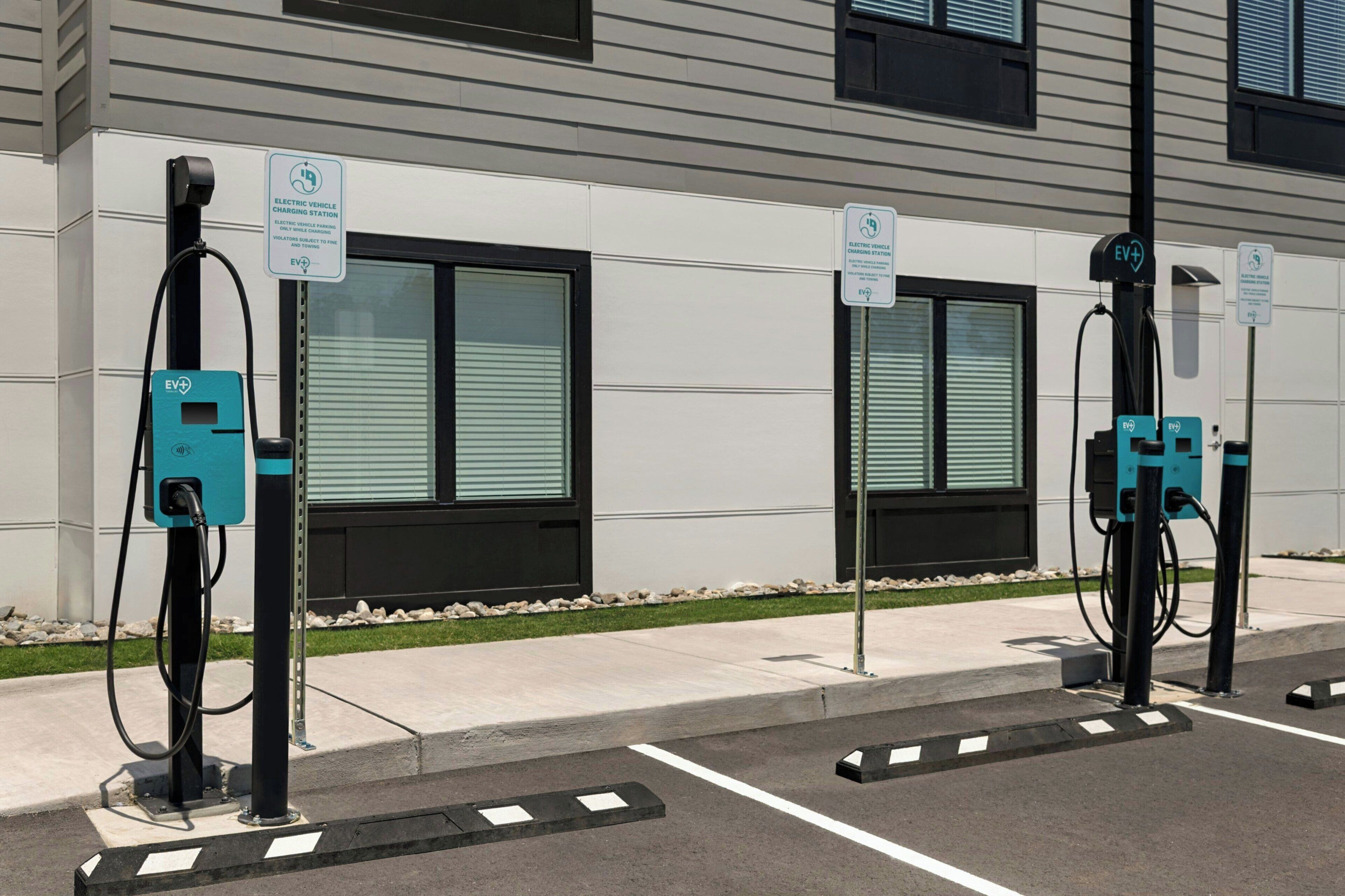 An electric vehicle charging station with two charging points, a sign indicating EV charging availability, and a building with large windows in the background.