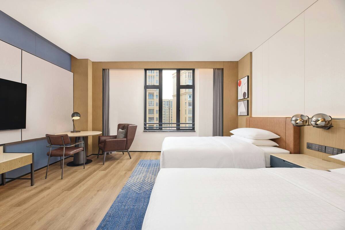 Habitación de hotel con dos camas, un escritorio, una silla, un televisor y un ventanal grande con vista a la ciudad.