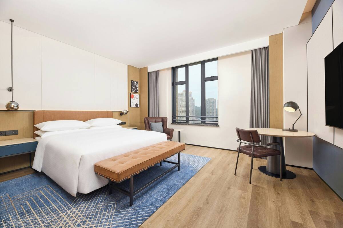 Una habitación de hotel con una cama grande, un escritorio, una silla, un banco, un televisor y una ventana con vista a la ciudad.