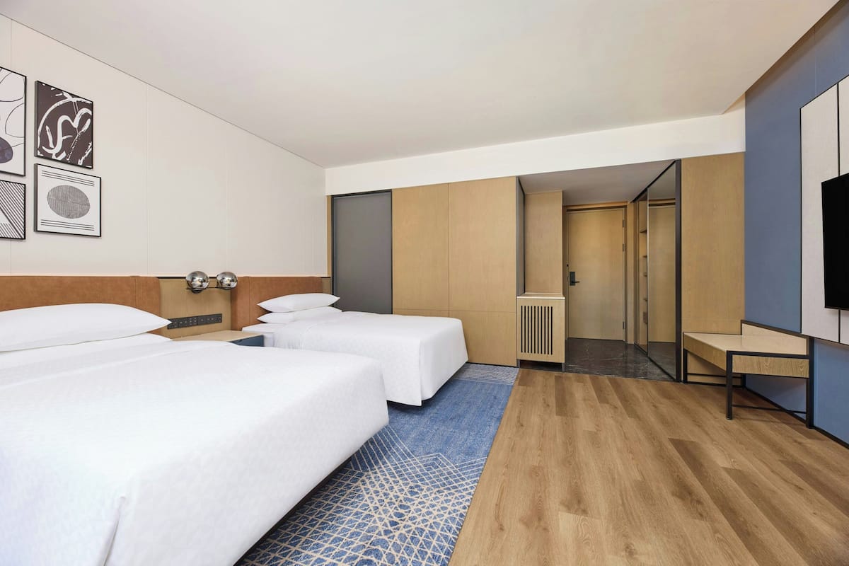 Habitación de hotel con dos camas, televisor, escritorio y baño visible a través de una puerta.