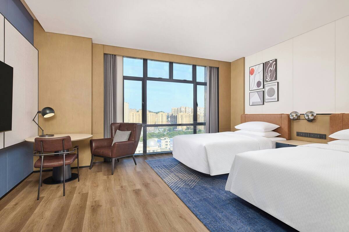 Habitación de hotel con dos camas, un escritorio, una silla, un televisor y un ventanal con vistas a la ciudad.