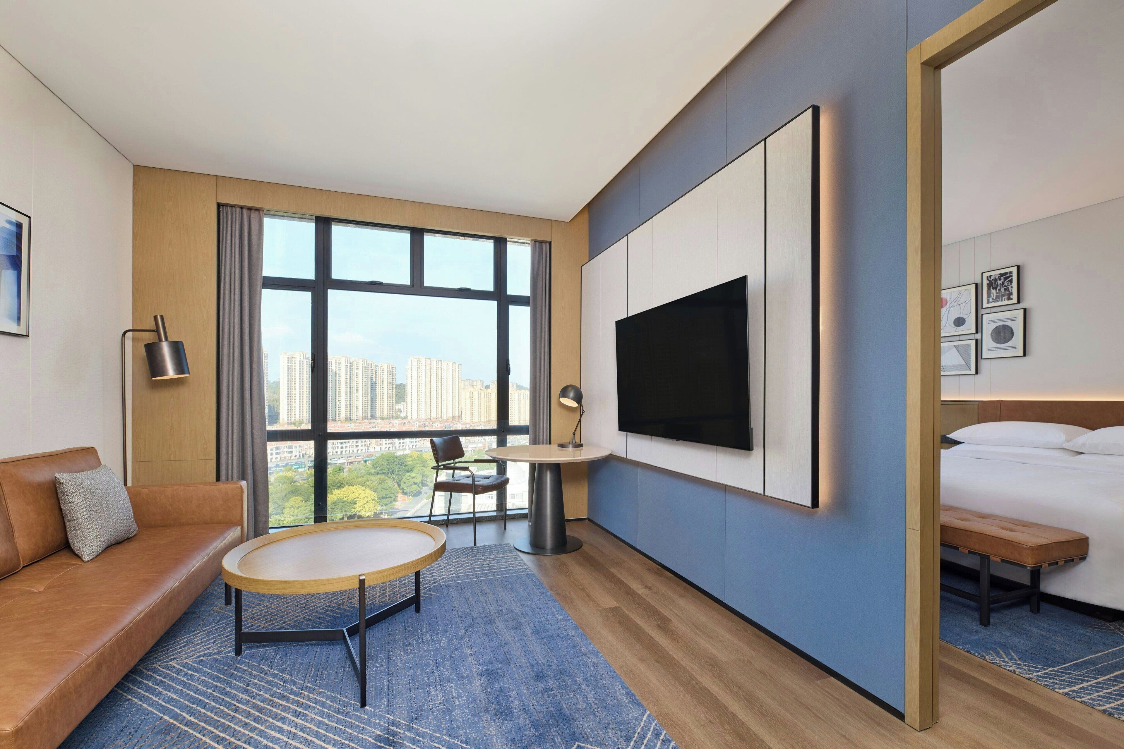 A modern hotel room with a large window, a sofa, a round table, a bed, and a TV mounted on the wall.