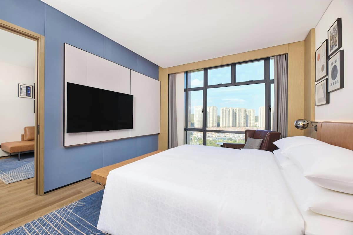 Una habitación de hotel moderna con una cama grande, un televisor montado en la pared, vistas a la ciudad y una zona de estar con una silla y una mesita.