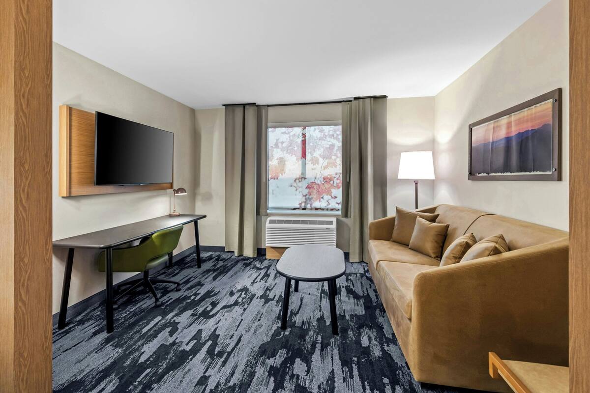A hotel room with a flat-screen TV, a sofa, a desk with a chair, a small table, and a window with curtains.