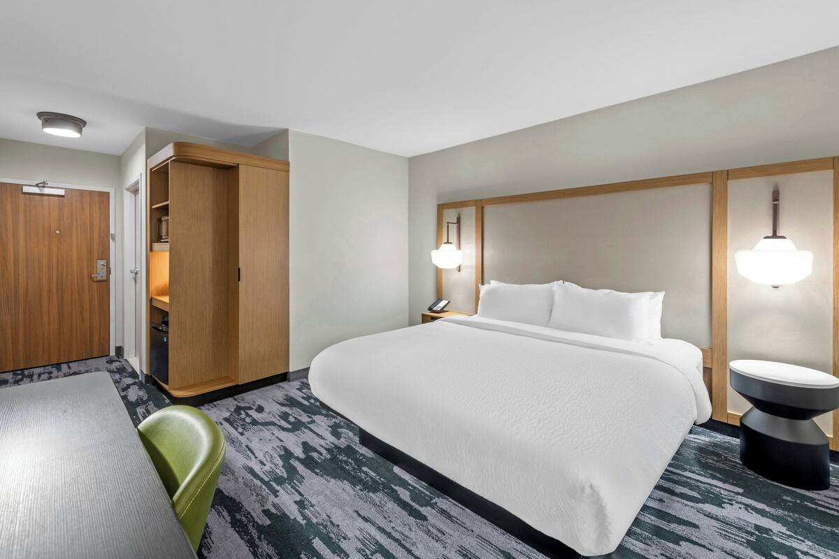 A hotel room with a large bed, a wooden wardrobe, a desk with a chair, and a bedside table with a lamp.