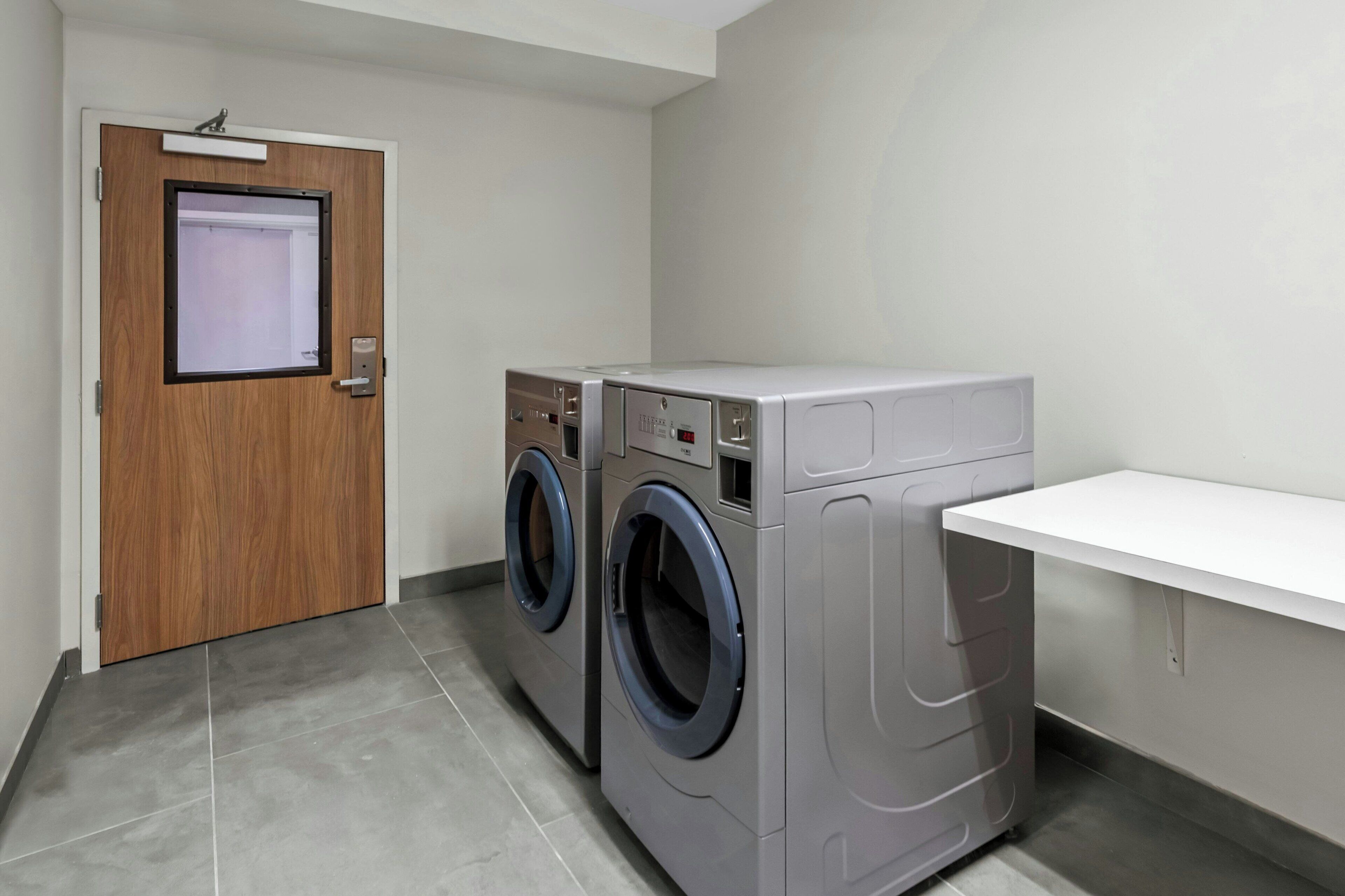 A laundry room with two front-loading washing machines, a wooden door with a window, and a white shelf.
