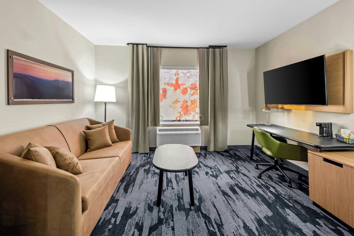 A hotel room with a sofa, a desk, a chair, a TV, a lamp, and a window with curtains.
