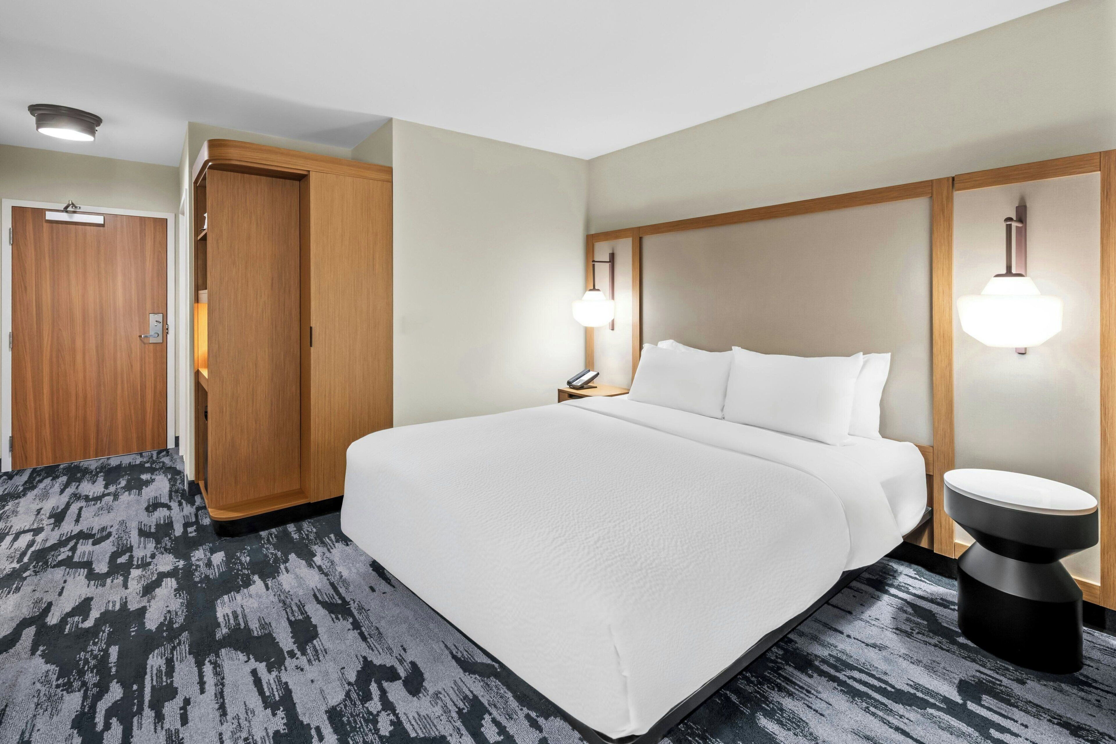A hotel room with a large bed, a wooden wardrobe, a bedside table with a lamp, and a carpeted floor.