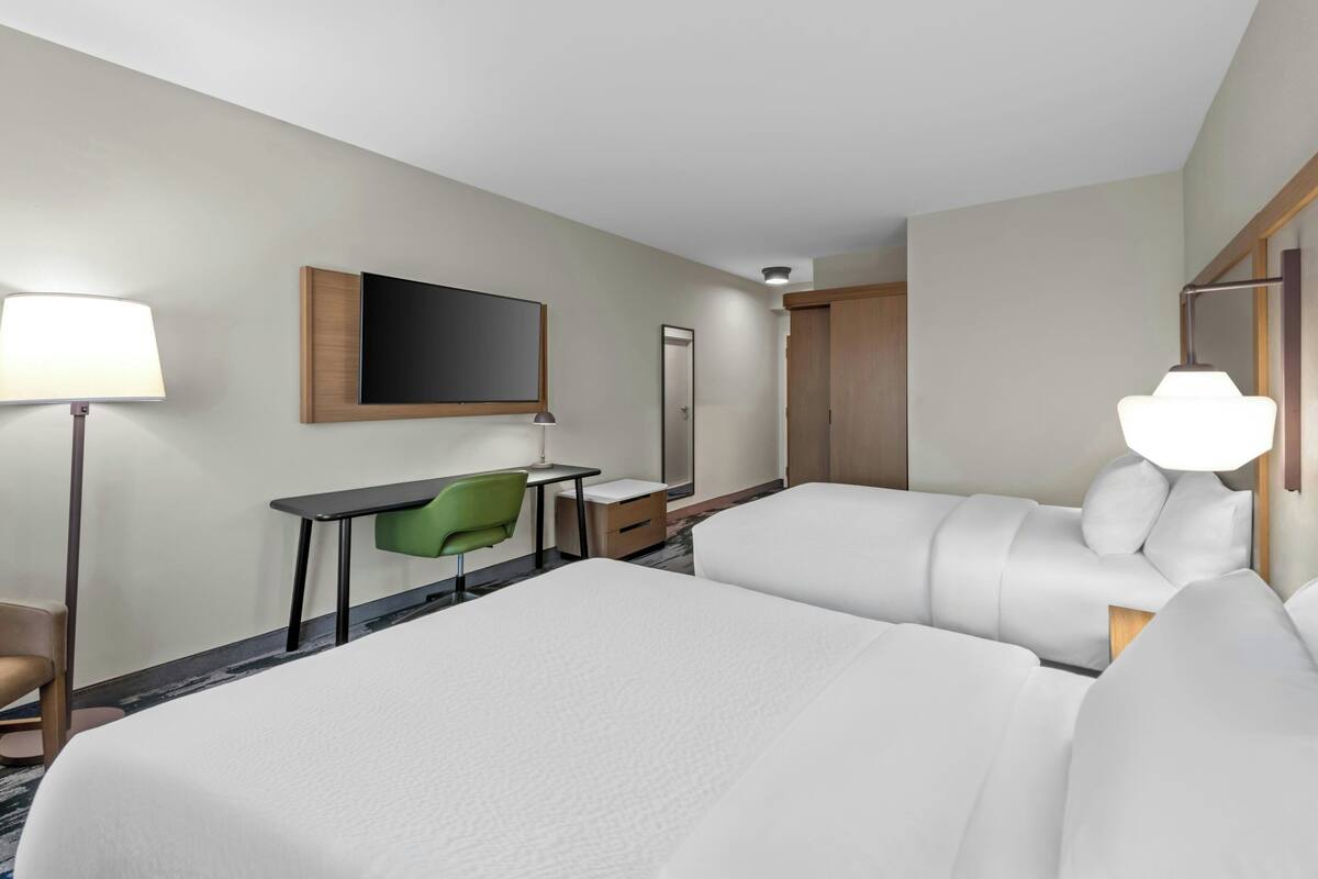 A hotel room with two beds, a desk, a chair, a TV, and a lamp.