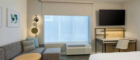A hotel room with a sofa, a small round table, a desk with a chair, a TV, and a large window.