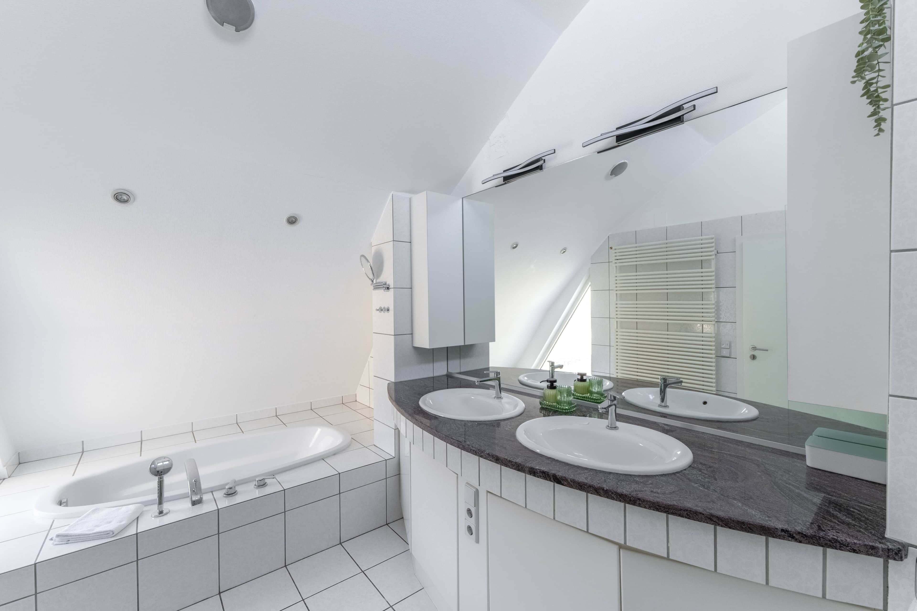 A modern bathroom with a bathtub, two sinks, a large mirror, and a plant.