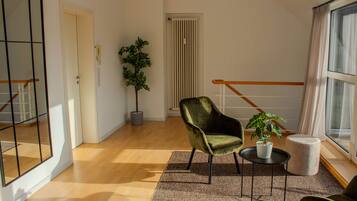 A spacious living room with a wooden floor, a green armchair, a small round table, a potted plant, and a large mirror.
