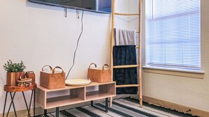 Une pièce avec un téléviseur fixé au mur, une échelle en bois, une fenêtre avec des stores, un tapis à rayures et une petite table sur laquelle se trouvent une plante et un sac.