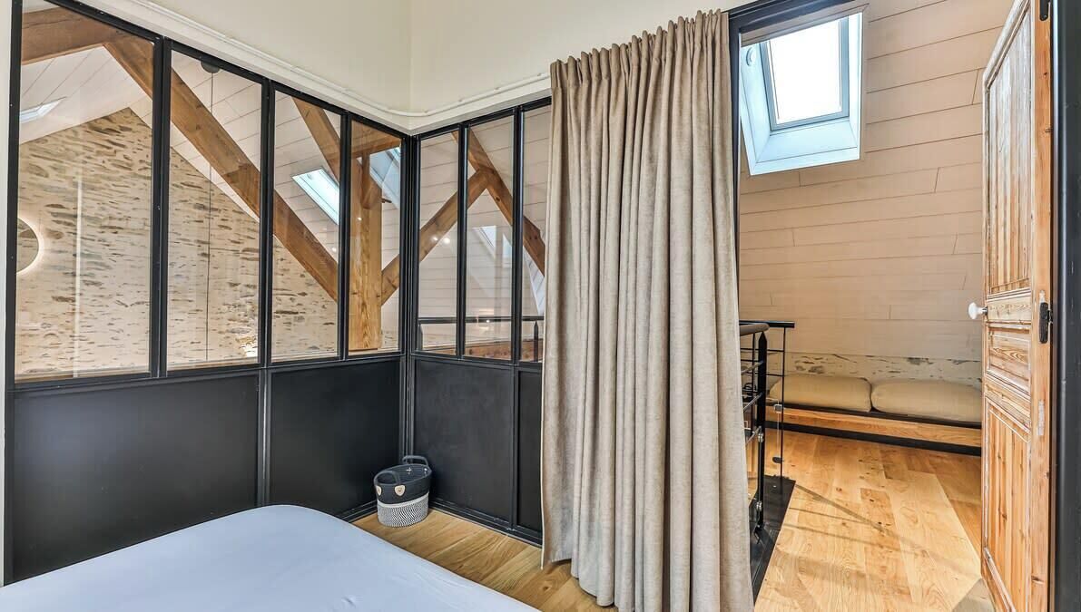 Een slaapkamer met een schuine kap, een bed, een raam met gordijnen en een zithoekje met kussens.