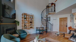 A spacious living room with a fireplace, a wooden bookshelf, a dining area, and a spiral staircase. - Warm & Cozy Condo • Prime Angel Fire Location (Angel Fire)