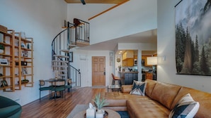 A spacious living room with a curved staircase, a wooden bookshelf, a leather sofa, and a dining area. - Warm & Cozy Condo • Prime Angel Fire Location (Angel Fire)