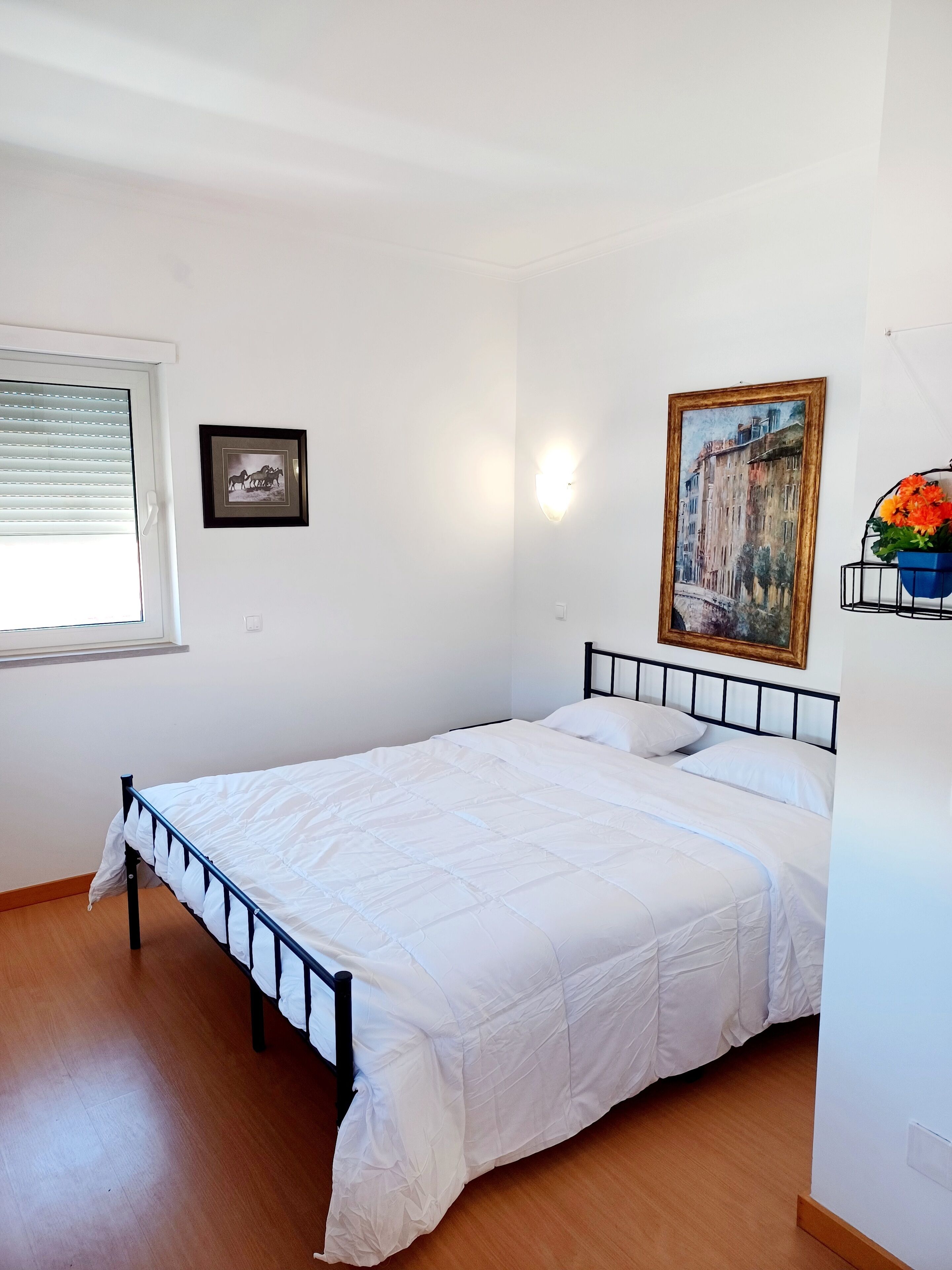 A bedroom with a bed, a window with blinds, a framed picture, a wall-mounted light, and a flower arrangement.