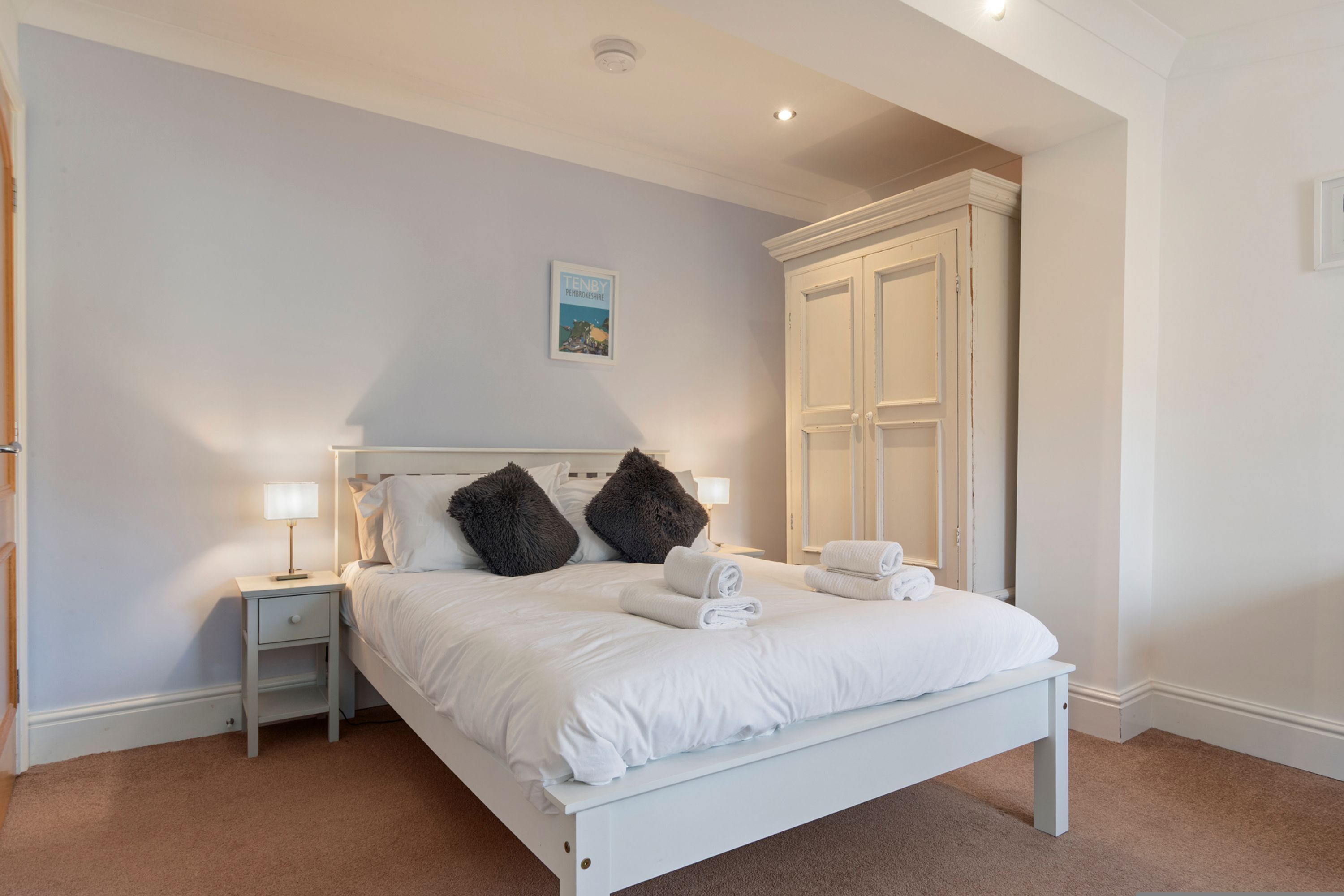 A neatly made bed with white linens and two dark pillows, a bedside table with a lamp, and a large white wardrobe.