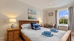 A neatly made bed with white linens and blue pillows, a wooden nightstand with a lamp, a desk with a chair, and a painting on the wall. - Daisy Cottage - St Florence Parking (Tenby)
