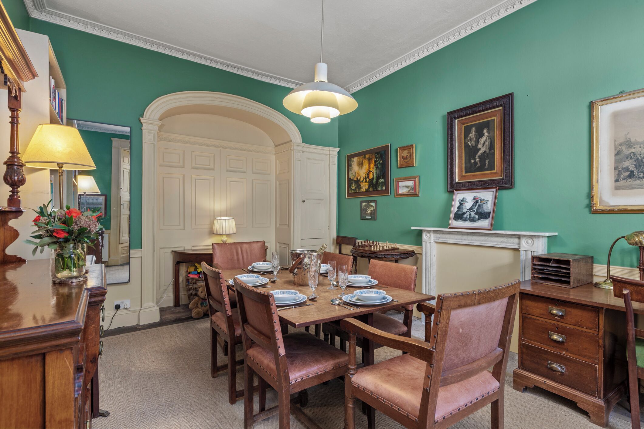 A dining room with a wooden table set for four, surrounded by upholstered chairs. A white door and a fireplace are visible in the background.