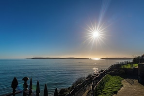 A coastal view with a clear sky, a sun setting or rising over the water, and a promenade with a fence and greenery. - St Mary s Seaview - In Town Walls Close to Beach (Tenby)