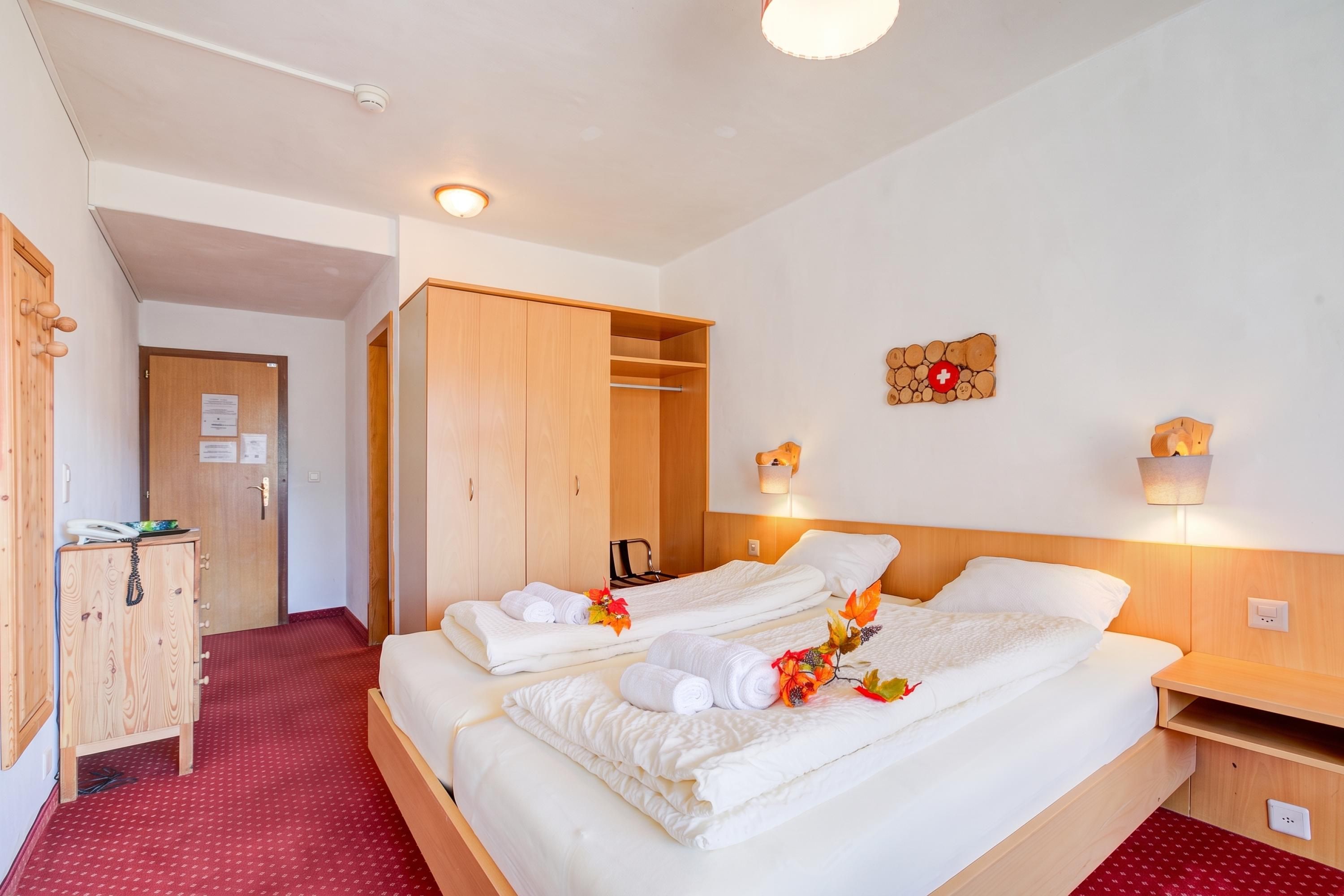 A hotel room with two beds, a wardrobe, a desk, and a wall-mounted light fixture.
