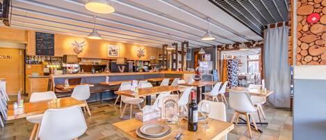 A restaurant interior with wooden tables, white chairs, hanging pendant lights, and a bar area with a menu board.