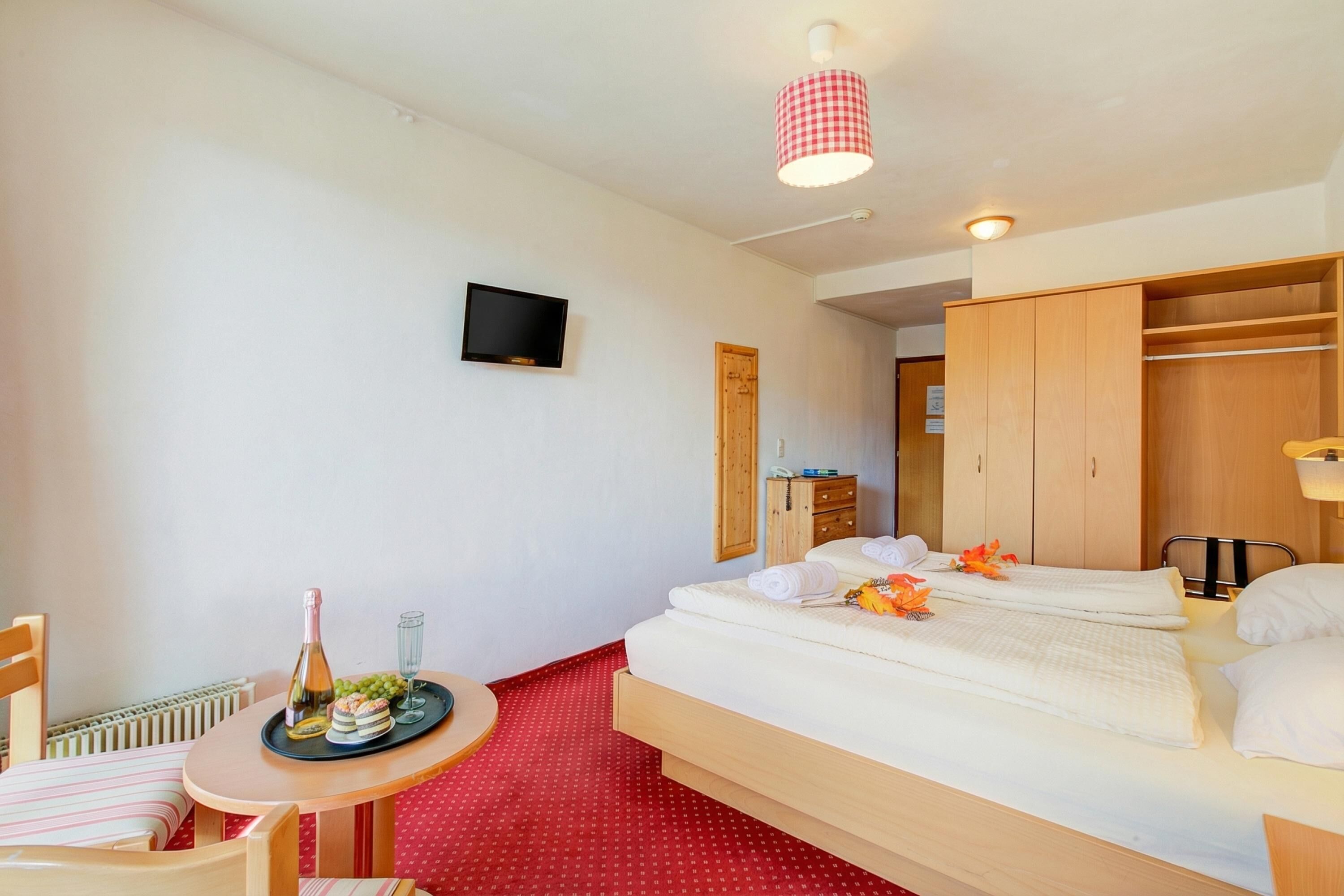 Ein Hotelzimmer mit einem Bett, einem kleinen Tisch mit Flasche und Gläsern, einem an der Wand montierten Fernseher und einem Holzschrank.