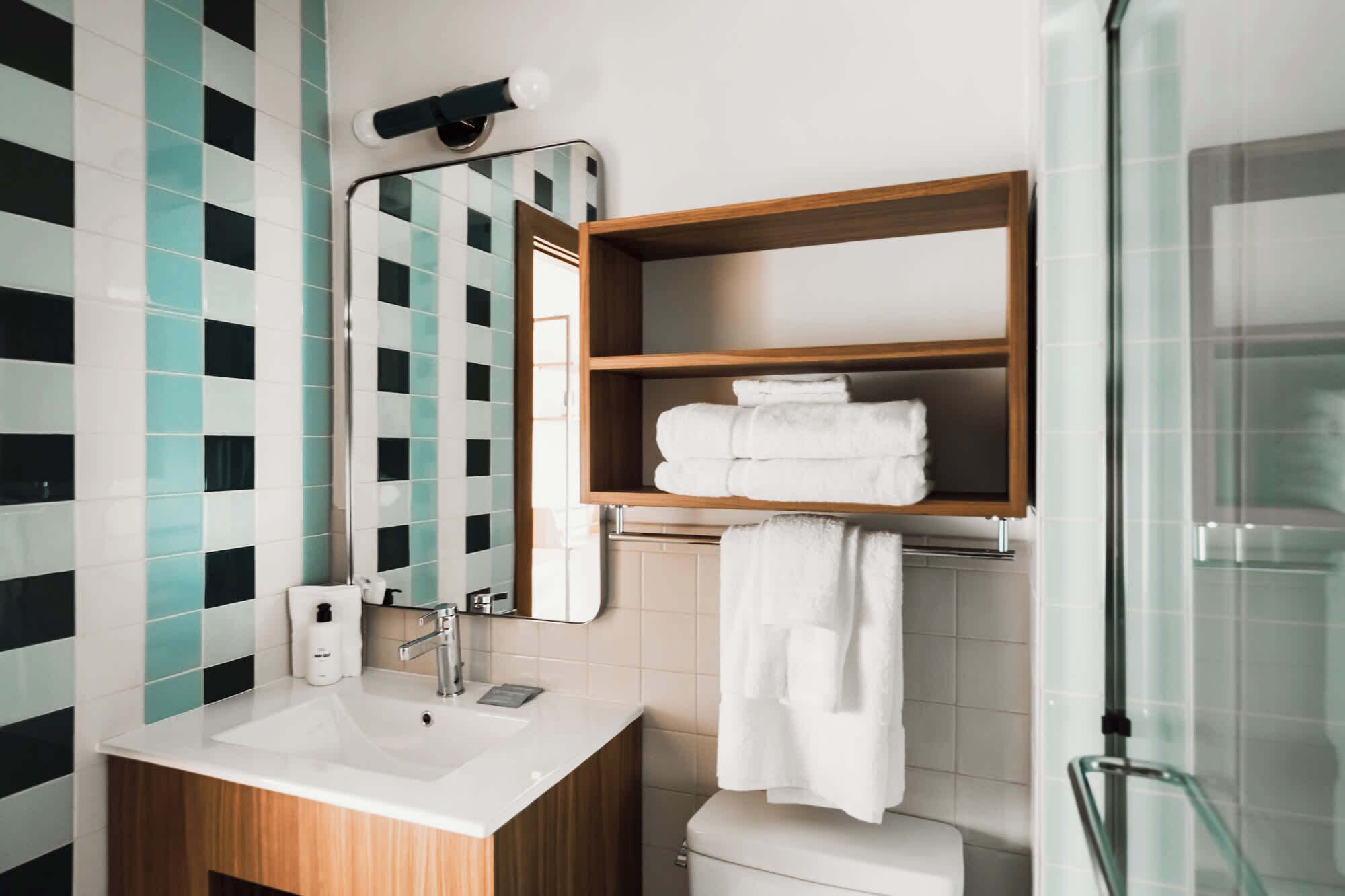 A modern bathroom with a white sink, a mirror, a wooden shelf with folded towels, and a glass shower enclosure.