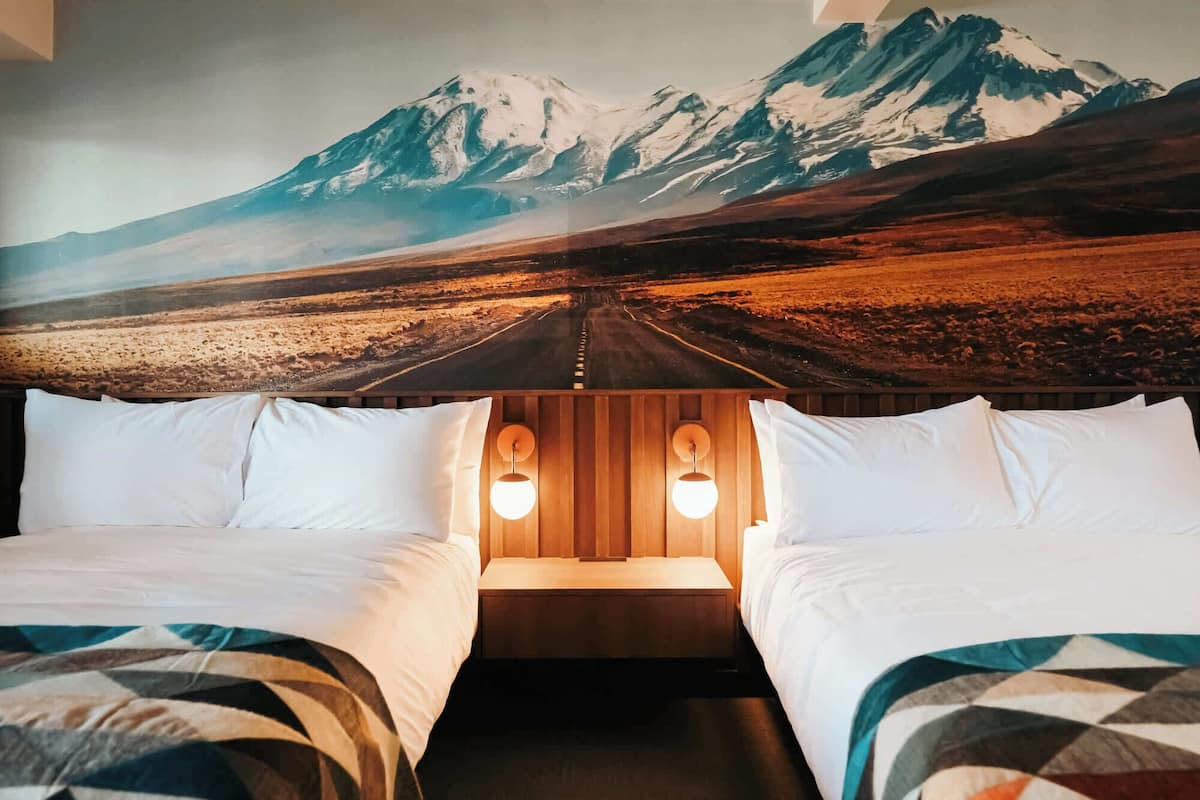 Habitación de hotel con dos camas, cabecera de madera y un mural grande con paisaje de montaña.