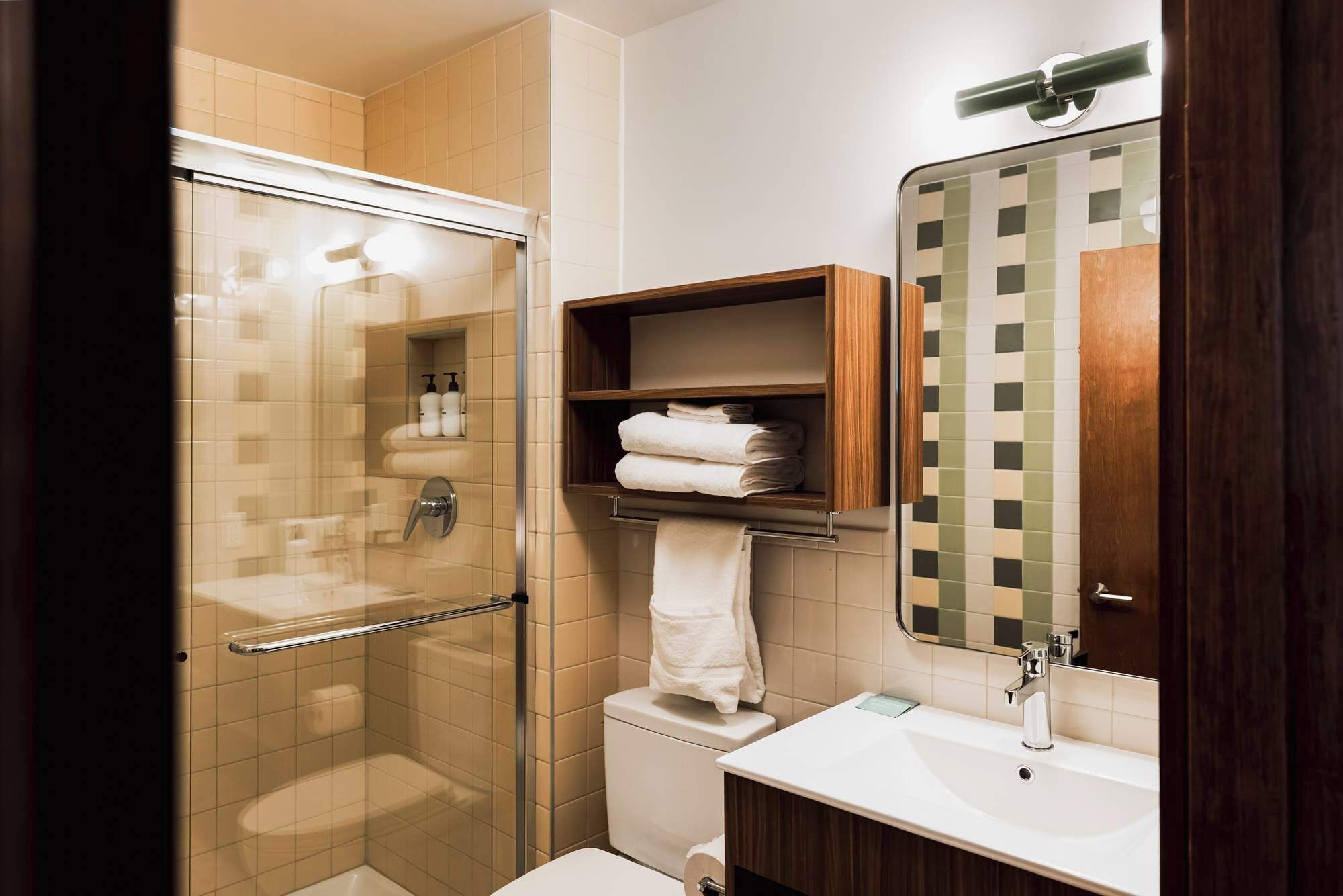 Une salle de bain avec une douche à parois de verre, des toilettes, un lavabo et une étagère en bois où sont posées des serviettes.