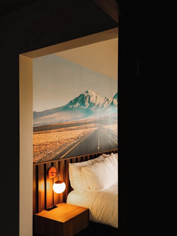 A bedroom with a large wall mural of a mountain road, a wooden headboard, a bedside table with a lamp, and a bed with white bedding.
