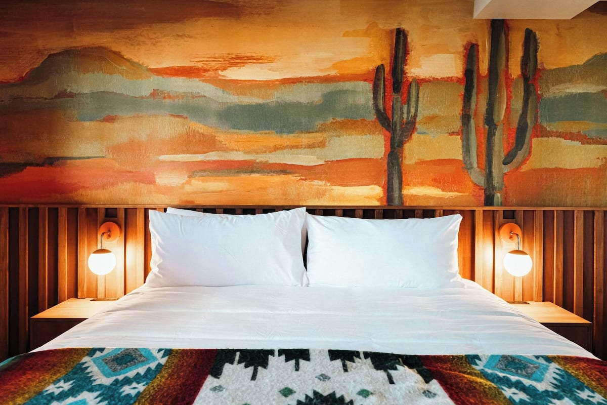 Una habitación de hotel con una cama, dos lámparas de noche, un tapete colorido y un cuadro grande de un paisaje desértico en la pared.